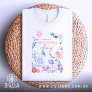 Футболка квіти прекрасний день, складена футболка на плетеному кошику, фон світло-сірий, принт з квітами та метеликами в пастельних кольорах, текст "Today is a BEAUTIFUL day" розташований в центрі.