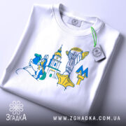 Футболка Київ білого кольору, складена на плоскій поверхні, фон світло-сірий, принт містить стилізовані зображення київських пам'яток, на футболці є етикетка з зеленим шнурком.