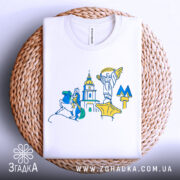 Футболка Київ білого кольору, складена на плоскій поверхні, фон білий, принт містить стилізовані зображення київських пам'яток, розташована на плетеній основі.