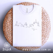 Футболка київський контур міста, біла, складена на плетеній кошику, фон світло-сірий, принт у вигляді контурів міста виконаний чорним кольором.