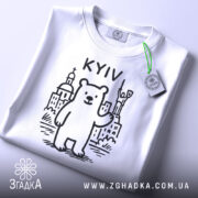 Футболка київський ведмідь, біла, складена на поверхні, фон світло-сірий, принт з ведмедем та архітектурними елементами Києва, напис 'KYIV' над ведмедем, на футболці є етикетка з зеленим шнурком.