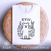 Футболка київський ведмідь, біла, складена на плетеній кошику, фон світло-сірий, принт з ведмедем та архітектурними елементами Києва, напис 'KYIV' над ведмедем.