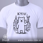 Футболка київський ведмідь, біла, складена на поверхні, фон світло-сірий, принт з ведмедем та архітектурними елементами Києва, напис 'KYIV' над ведмедем, на футболці є етикетка з зеленим шнурком.