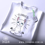 Футболка лама Бердичів, біла, складена на столі, фон світло-сірий, принт з лами в позі медитації з написом 'LLAMASTE', на футболці є етикетка з зеленим шнурком.