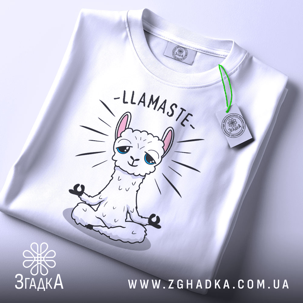 Футболка лама Бердичів, біла, складена на столі, фон світло-сірий, принт з лами в позі медитації з написом 'LLAMASTE', на футболці є етикетка з зеленим шнурком.