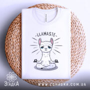 Футболка лама Бердичів, біла, складена на плетеному кошику, фон світло-сірий, принт з лами в позі медитації з написом 'LLAMASTE'.