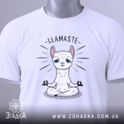 Футболка лама Бердичів, біла, складена на столі, фон світло-сірий, принт з лами в позі медитації з написом 'LLAMASTE', на футболці є етикетка з зеленим шнурком.