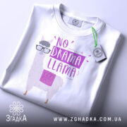 Футболка лама без драми, біла, складена на поверхні, фон світло-сірий, принт з лемуром у окулярах та написом 'NO DRAMA LLAMA' у фіолетових тонах, з етикеткою на комірці.