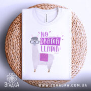 Футболка лама без драми, біла, складена на плетеній кошику, фон світло-сірий, принт з лемуром у окулярах та написом 'NO DRAMA LLAMA' у фіолетових тонах.