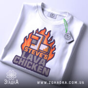 Футболка лава курка Майкрафт, біла, лежить на столі, фон світло-сірий, принт з вогняними елементами та текстом 'STEVE'S LAVA CHICKEN' у фіолетових та помаранчевих кольорах, з етикеткою на зеленому шнурку.