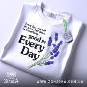 Футболка лаванда гарний настрій. Біла футболка, складена на поверхні, з принтом, що містить текст і зображення лаванди. Принт розташований по центру, текст чорного кольору, а лаванда виконана в фіолетових відтінках. На футболці є етикетка з зеленим шнурком, фон світло-сірий.