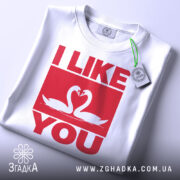 Футболка лебеді кохання, біла, складена на поверхні, з видимим етикеткою, фон світло-сірий, принт червоного кольору з написом 'I LIKE YOU' та двома лебедями, що з'єднують свої голови.