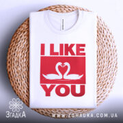 Футболка лебеді кохання, біла, складена на плетеній кошику, фон світло-сірий, принт червоного кольору з написом 'I LIKE YOU' та двома лебедями, що з'єднують свої голови.
