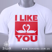 Футболка лебеді кохання, біла, складена на поверхні, з видимим етикеткою, фон світло-сірий, принт червоного кольору з написом 'I LIKE YOU' та двома лебедями, що з'єднують свої голови.