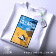 Футболка лелеки летять, біла, зображена на столі, фон світло-сірий, принт з лелекою на жовтому полі, з етикеткою на зеленій стрічці.