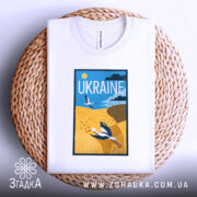 Футболка лелеки летять, біла, зображена на круглому кошику, фон світло-сірий, принт з лелекою на жовтому полі та написом 'UKRAINE' у верхній частині.