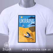 Футболка лелеки летять, біла, зображена на столі, фон світло-сірий, принт з лелекою на жовтому полі, з етикеткою на зеленій стрічці.