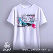 Футболка Леся емоції. Біла футболка, підвішена на вішалці, з принтом, що зображує жінку з коротким волоссям. На фоні кольорові плями та текст українською мовою.