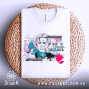 Футболка Леся емоції. Біла футболка, складена на плетеній основі, з принтом, що зображує жінку з коротким волоссям. Принт містить кольорові елементи та текст українською мовою.