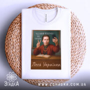 Футболка Леся Українка білого кольору, зображена на плетеній основі, принт з портретом жінки з телефоном, яка показує жест миру, на фоні дошки з написом 'Чесний покрив не потрібен'.