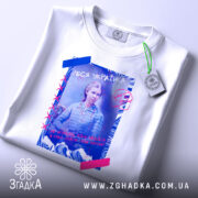 Футболка Леся Українка білого кольору, зображена з верхнього ракурсу, на фоні світло-сірого кольору, принт з Лесею Українкою, з кольоровими елементами, на футболці є етикетка з зеленим шнурком.