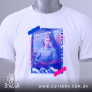 Футболка Леся Українка білого кольору, зображена з верхнього ракурсу, на фоні світло-сірого кольору, принт з Лесею Українкою, з кольоровими елементами, на футболці є етикетка з зеленим шнурком.