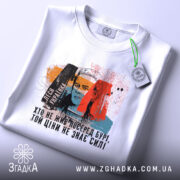 Футболка Леся Українка, білого кольору, зображена зверху, на столі, фон світлий, принт з портретом Лесі Українки та кольоровими елементами, на футболці є етикетка.