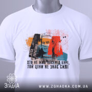 Футболка Леся Українка, білого кольору, зображена зверху, на столі, фон світлий, принт з портретом Лесі Українки та кольоровими елементами, на футболці є етикетка.