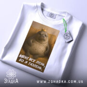 Футболка лєнь я тюлень, складена біла футболка з принтом, зображення тюленя з короною на фоні жовтого дивану, текст на футболці: 'МЕНІ ВСЕ ЛЄНЬ, БО Я ТЮЛЕНЬ', на футболці є етикетка.