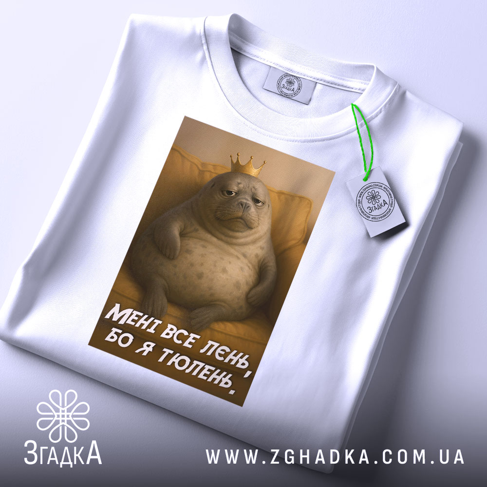 Футболка лєнь я тюлень, складена біла футболка з принтом, зображення тюленя з короною на фоні жовтого дивану, текст на футболці: 'МЕНІ ВСЕ ЛЄНЬ, БО Я ТЮЛЕНЬ', на футболці є етикетка.