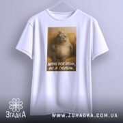 Футболка лєнь я тюлень, біла футболка з принтом, зображення тюленя з короною на фоні жовтого дивану, текст на футболці: 'МЕНІ ВСЕ ЛЄНЬ, БО Я ТЮЛЕНЬ', футболка підвішена на вішалці.