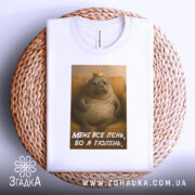 Футболка лєнь я тюлень, складена біла футболка з принтом, зображення тюленя з короною на фоні жовтого дивану, текст на футболці: 'МЕНІ ВСЕ ЛЄНЬ, БО Я ТЮЛЕНЬ', футболка лежить на плетеній основі.