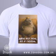 Футболка лєнь я тюлень, складена біла футболка з принтом, зображення тюленя з короною на фоні жовтого дивану, текст на футболці: 'МЕНІ ВСЕ ЛЄНЬ, БО Я ТЮЛЕНЬ', на футболці є етикетка.