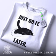 Футболка лінивий кіт, білого кольору, складена на поверхні, фон світло-сірий, принт чорного кота, що лежить на логотипі, з текстом 'JUST DO IT. LATER.' у чорному кольорі, на комірці видна етикетка.