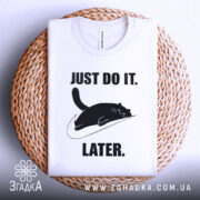 Футболка лінивий кіт, білого кольору, складена на плетеній кошику, фон світло-сірий, принт чорного кота, що лежить на логотипі, з текстом 'JUST DO IT. LATER.' у чорному кольорі.