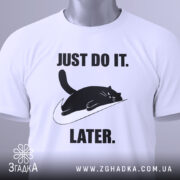 Футболка лінивий кіт, білого кольору, складена на поверхні, фон світло-сірий, принт чорного кота, що лежить на логотипі, з текстом 'JUST DO IT. LATER.' у чорному кольорі, на комірці видна етикетка.