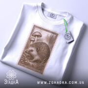 Футболка лісовий їжачок білого кольору, складена на поверхні, ракурс зверху, фон світло-сірий, принт з їжачком та грибами, прикріплена етикетка з логотипом.