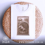 Футболка лісовий їжачок білого кольору, складена на плетеній кошику, ракурс зверху, фон світло-сірий, принт з їжачком та грибами, видима етикетка з логотипом.