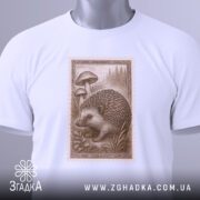 Футболка лісовий їжачок білого кольору, складена на поверхні, ракурс зверху, фон світло-сірий, принт з їжачком та грибами, прикріплена етикетка з логотипом.