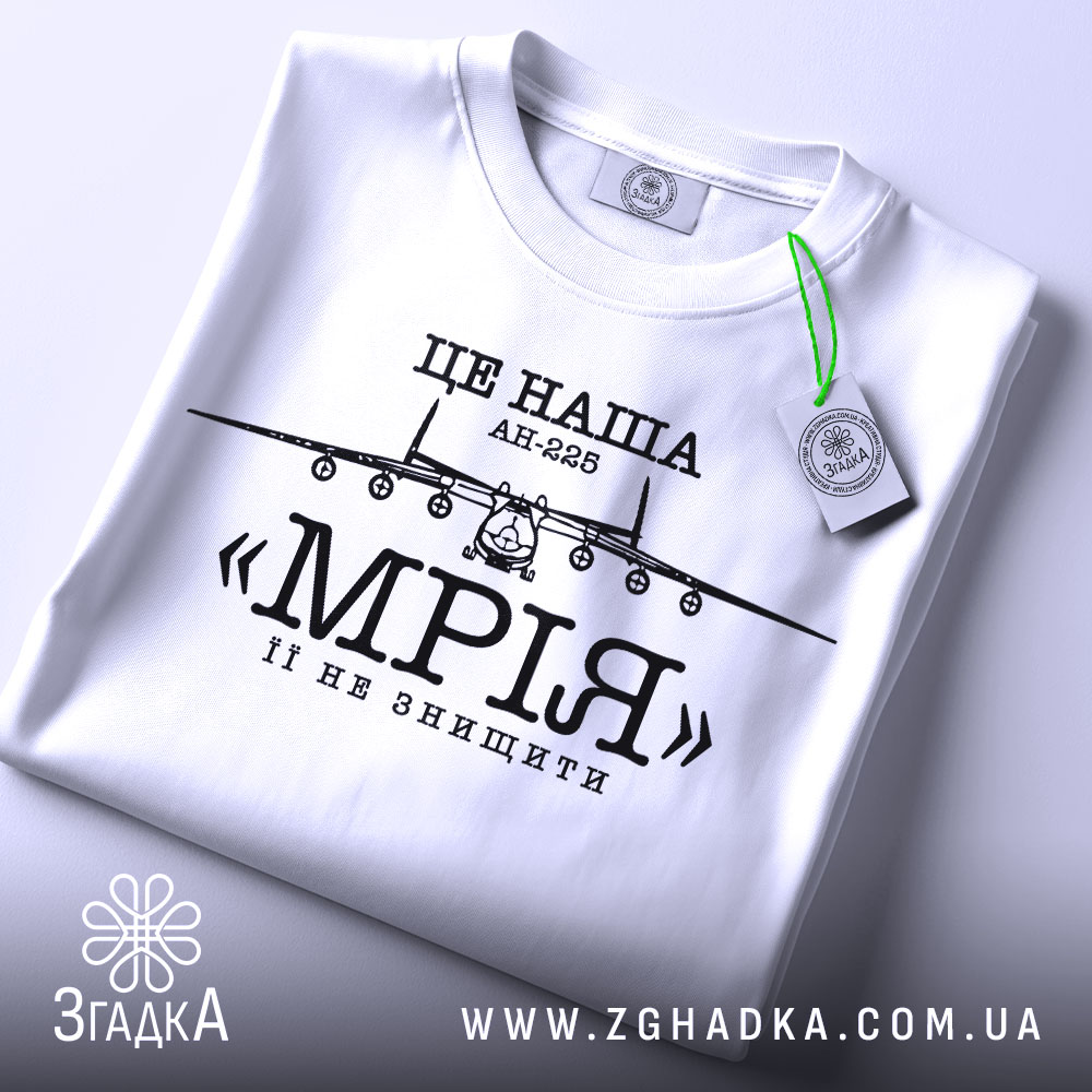 Футболка літак мрія, біла, складена на столі, з видимим етикеткою, фон світлий, принт з літаком АН-225 та написом «МРІЯ» чорного кольору.