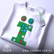 Футболка літера ї, біла, зображена зверху, фон світлий, принт у вигляді зеленого листя з червоними та синіми квітами, на футболці є етикетка.