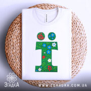 Футболка літера ї, біла, складена на плетеній кошику, фон світлий, принт у вигляді зеленого листя з червоними та синіми квітами.