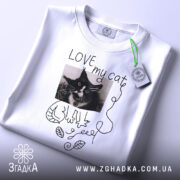 Футболка люблю мого кота, білого кольору, складена на поверхні, фон світло-сірий, принт з чорно-білим зображенням кота, напис 'LOVE my cat' та зелений ярлик.