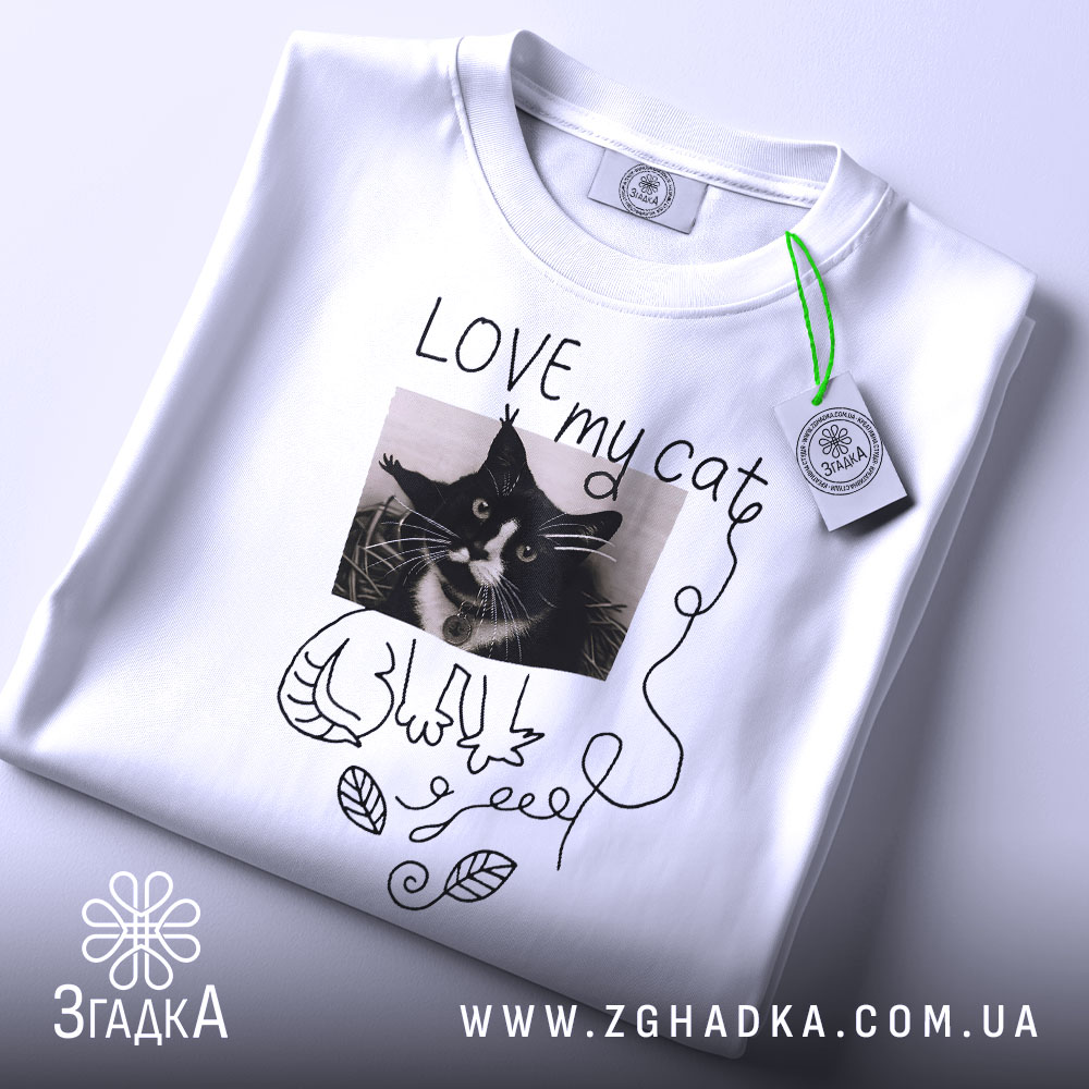 Футболка люблю мого кота, білого кольору, складена на поверхні, фон світло-сірий, принт з чорно-білим зображенням кота, напис 'LOVE my cat' та зелений ярлик.