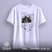Футболка люблю мого кота, білого кольору, підвішена на вішалці, фон світло-сірий, принт з чорно-білим зображенням кота та написом 'LOVE my cat' у рукописному стилі.