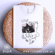 Футболка люблю мого кота, білого кольору, складена на плетеній кошику, фон білий, принт з чорно-білим зображенням кота, напис 'LOVE my cat' та деталі у вигляді листя.