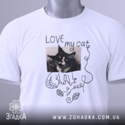Футболка люблю мого кота, білого кольору, складена на поверхні, фон світло-сірий, принт з чорно-білим зображенням кота, напис 'LOVE my cat' та зелений ярлик.