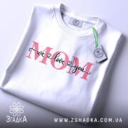 Футболка любов до мами, біла, зображена зверху, фон світло-сірий, принт з написом 'MOM' червоного кольору та 'we love you' чорного кольору, з етикеткою зеленого кольору.