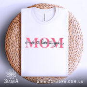 Футболка любов до мами, біла, зображена зверху на плетеній кошику, фон світло-сірий, принт з написом 'MOM' червоного кольору та 'we love you' чорного кольору.