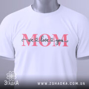 Футболка любов до мами, біла, зображена зверху, фон світло-сірий, принт з написом 'MOM' червоного кольору та 'we love you' чорного кольору, з етикеткою зеленого кольору.